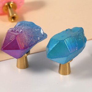 Peut inclure: Gros plan sur des boutons de meubles d&eacute;coratifs avec des formations cristallines. Les boutons pr&eacute;sentent une vari&eacute;t&eacute; de couleurs, dont le bleu, le violet et le rose, avec des bases dor&eacute;es. Ces boutons uniques ajoutent une touche d'&eacute;l&eacute;gance.