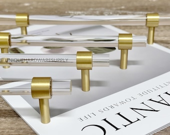 May include: A set of four clear acrylic cabinet pulls with brushed brass metal posts. Each handle features a transparent cylindrical bar secured by two metallic gold-toned supports. The text UNIQUEHARDWARESUPPLY is printed on one of the acrylic bars.