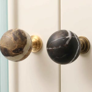 May include: Two spherical cabinet knobs mounted on a cream-colored surface. The left knob is made of brown and beige marbled stone, while the right knob is black with white veining. Both knobs feature a circular brass-toned metal base attachment.