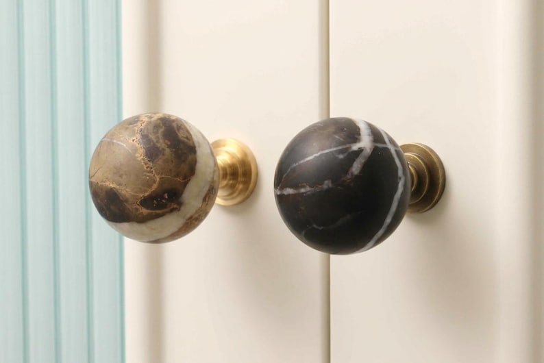 May include: Two spherical cabinet knobs mounted on a cream-colored surface. The left knob is made of brown and beige marbled stone, while the right knob is black with white veining. Both knobs feature a circular brass-toned metal base attachment.