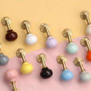 Op de afbeelding: Een collectie van twaalf hangende kastgrepen met gladde, ronde keramische knoppen in diverse kleuren, waaronder wit, zwart, bruin, geel, oranje, roze, lichtblauw, lavendel en grijs, bevestigd aan beslag van geborsteld goudkleurig metaal.
