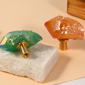 Peut inclure: Deux boutons de meuble en cristal. L'un est vert avec une base dor&eacute;e, l'autre est orange avec une base dor&eacute;e. Les boutons sont pos&eacute;s sur une pierre blanche et une surface couleur p&ecirc;che. Les boutons sont d&eacute;coratifs.