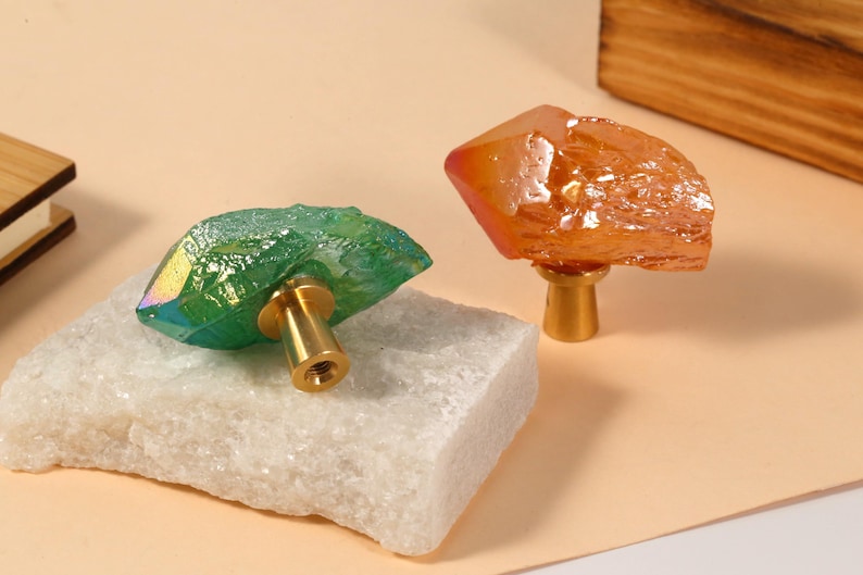 Peut inclure: Deux boutons de meuble en cristal. L'un est vert avec une base dor&eacute;e, l'autre est orange avec une base dor&eacute;e. Les boutons sont pos&eacute;s sur une pierre blanche et une surface couleur p&ecirc;che. Les boutons sont d&eacute;coratifs.