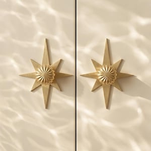 Op de afbeelding: Twee goudkleurige metalen kastknoppen gemonteerd op een lichtbeige oppervlak. Elke knop heeft een centraal rond geribbeld element omringd door een platte, achtpuntige stervormige achterplaat met scherpe, langwerpige stralen.