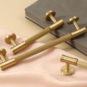 May include: A collection of brushed gold cabinet pulls and knobs featuring a cylindrical design with vertical ribbed detailing. Each piece includes circular mounting bases and a metallic finish, showcasing a consistent texture and hardware style.