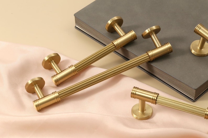 May include: A collection of brushed gold cabinet pulls and knobs featuring a cylindrical design with vertical ribbed detailing. Each piece includes circular mounting bases and a metallic finish, showcasing a consistent texture and hardware style.