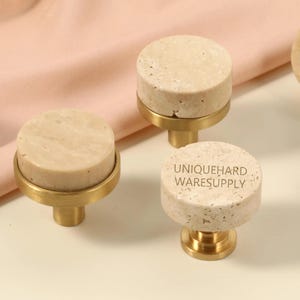 May include: A collection of cabinet knobs with a light beige, textured stone top and a gold-colored base. The knobs are cylindrical and have the text "UNIQUEHARD WARESUPPLY" printed on one of them. The background is a light pink fabric.