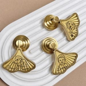 May include: Three brass cabinet pulls with a fan-shaped design. One pull features a dog portrait with the name "Max" and a paw print. Another shows a couple kissing with the names "David & Emma." The third pull has a child's portrait and the name "Lucas."