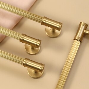 May include: A set of four gold-colored metal cabinet pulls featuring a cylindrical, fluted design with ribbed detailing at the ends and circular mounting bases. The hardware has a brushed finish and is arranged in a parallel formation.