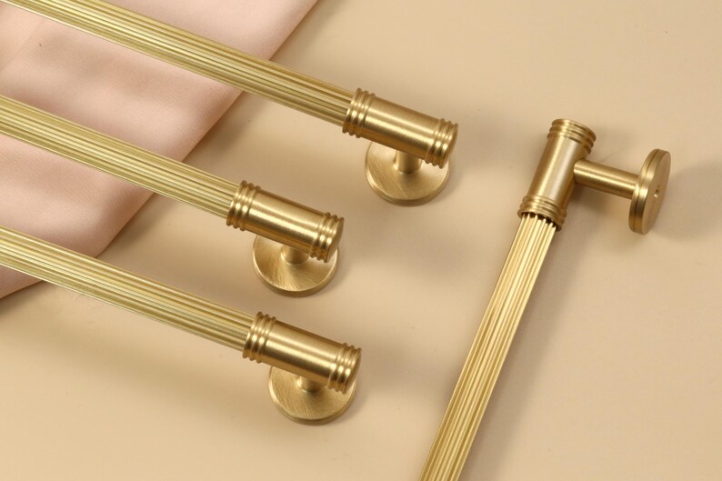 May include: A set of four gold-colored metal cabinet pulls featuring a cylindrical, fluted design with ribbed detailing at the ends and circular mounting bases. The hardware has a brushed finish and is arranged in a parallel formation.