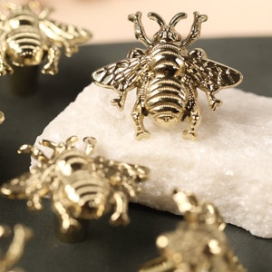 May include: Five gold bee-shaped drawer pulls on a dark gray surface. The bee pulls have a detailed design and are a popular choice for home decor.