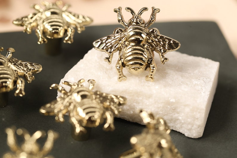 May include: Five gold bee-shaped drawer pulls on a dark gray surface. The bee pulls have a detailed design and are a popular choice for home decor.