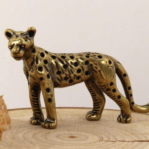 May include: A detailed, golden-colored leopard figurine with black spots. The sculpture is posed in a standing position, with its tail curved. The leopard is placed on a wooden surface, showcasing its intricate design and craftsmanship.