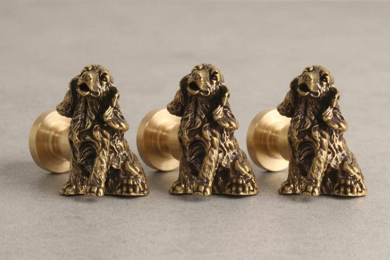 Peut inclure: Trois boutons de placard en laiton, chacun repr&eacute;sentant une sculpture tridimensionnelle d&eacute;taill&eacute;e d'un &eacute;pagneul assis avec de longues oreilles textur&eacute;es et une finition m&eacute;tallique. Les boutons ont une base cylindrique pour la fixation.