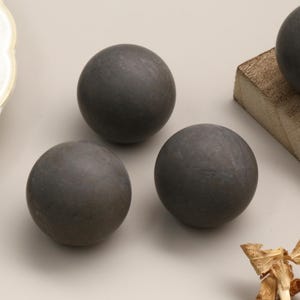 Op de afbeelding: Vier donkergrijze bollen, mogelijk decoratieve bollen, zijn gerangschikt op een lichtbeige oppervlak. Eén bol rust op een klein houten blok. De bollen hebben een textuur. Een decoratieve plaat is zichtbaar op de achtergrond.
