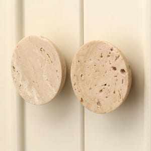 May include: Two circular cabinet knobs made of beige travertine stone with a natural porous texture and visible mineral veining. The knobs have a smooth, rounded edge profile and are mounted side by side on a vertical paneled surface.