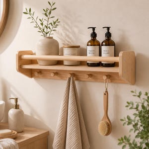 May include: A light wood bathroom shelf with a towel rack, holding a vase with greenery, a candle, and two brown glass bottles labeled "Hand Wash" and "Body Wash." A towel and a wooden brush hang below.