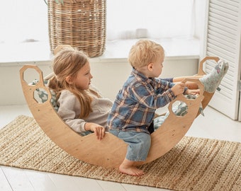 May include: Two children are playing on a wooden rocking balance board. The board is shaped like a crescent moon and has a honeycomb pattern on the sides. The children are sitting on the board and rocking back and forth. The girl is wearing a light brown sweater and the boy is wearing a blue and black plaid shirt.