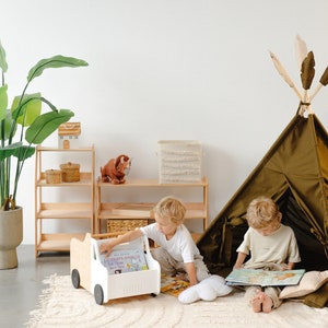 Puede incluir: Dos ni&ntilde;os est&aacute;n jugando en una sala de juegos. Un ni&ntilde;o est&aacute; sentado en una tienda de campa&ntilde;a tipi leyendo un libro. El otro ni&ntilde;o est&aacute; sentado en el suelo junto a un cami&oacute;n de juguete blanco con un libro dentro. El cami&oacute;n tiene una base de madera y ruedas blancas. La sala de juegos tiene una estanter&iacute;a de madera con un dinosaurio de juguete y una cesta encima.
