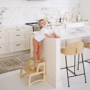 May include: A light-colored wooden step stool with two steps. The stool has a curved design and is placed in a kitchen setting. A child is standing on the stool, leaning on a white countertop. The kitchen features white cabinets and a marble countertop.