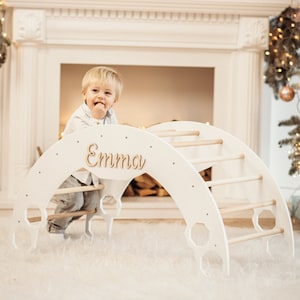 May include: A white wooden climbing arch with a personalized name plaque that reads "Emma". A young child is climbing on the arch. The arch is in a room decorated with a fireplace and Christmas decorations.