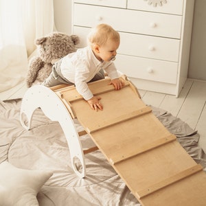 May include: A white wooden climbing arch with a natural wood ramp is in a child's playroom. A baby is climbing on the ramp. A white dresser with eyelash-shaped drawer pulls and a framed print that says "I love you more than all the stars" are in the background.