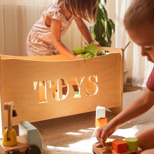 May include: A light-coloured wooden toy box with the word "TOYS" cut out on the front. Children are playing with toys, including wooden blocks and a toy truck. The toy box is approximately 61 cm long.