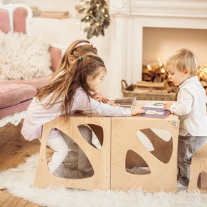 May include: Three children sitting at a wooden table with cutouts, reading a book together. The table is made of light wood and has a unique design. The children are sitting on stools and a step stool.