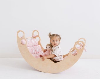 May include: A curved wooden rocker featuring a light-colored plywood frame with hexagonal cutouts at the ends. The interior is lined with a soft, tufted pink cushion decorated with a white pattern, providing a comfortable seat for play.