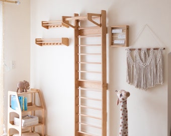 Puede incluir: Una escalera de escalada de madera con una barra en la parte superior para hacer dominadas, montada en una pared blanca. Hay dos estantes de madera encima de la escalera y una pared de macramé con las palabras "Love You More" en un letrero a la derecha de la escalera.