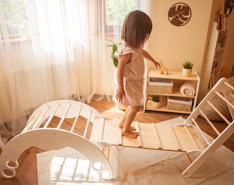 May include: A white wooden climbing frame with a ladder and a curved ramp. A young child is climbing on the ramp.