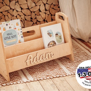 Puede incluir: Porta libros de madera con dos estantes y un nombre personalizado "Adam" en letra cursiva. El porta libros est&aacute; hecho de madera natural y se muestra en una alfombra marr&oacute;n y blanca.