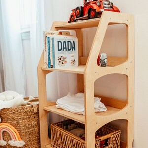 K&ouml;nnte beinhalten: Ein helles Holzregal mit drei Ebenen. Auf der obersten Ablage steht ein roter Spielzeuglaster. Die mittlere Ebene enth&auml;lt eine Reihe von Kinderb&uuml;chern, darunter eines mit dem Titel DADA von Jimmy Fallon, sowie ein gefaltetes wei&szlig;es Kleidungsst&uuml;ck.