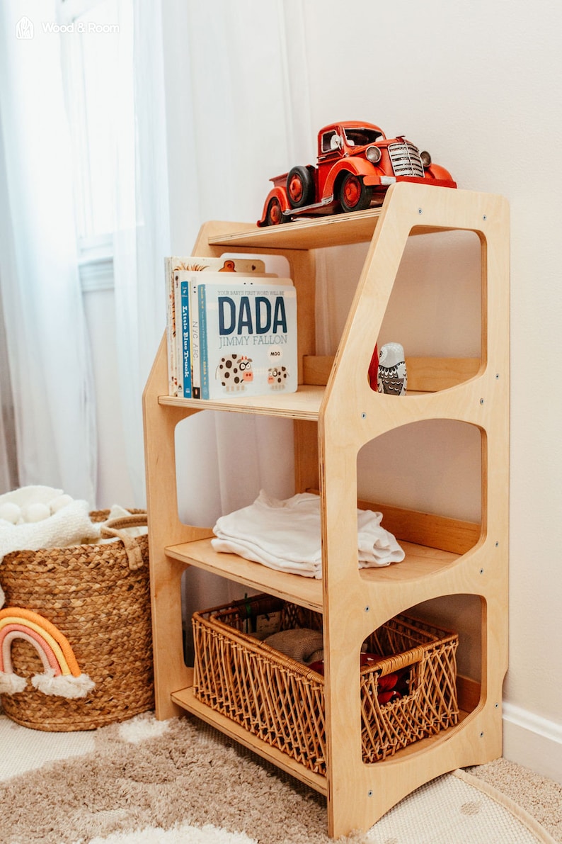 K&ouml;nnte beinhalten: Ein helles Holzregal mit drei Ebenen. Auf der obersten Ablage steht ein roter Spielzeuglaster. Die mittlere Ebene enth&auml;lt eine Reihe von Kinderb&uuml;chern, darunter eines mit dem Titel DADA von Jimmy Fallon, sowie ein gefaltetes wei&szlig;es Kleidungsst&uuml;ck.
