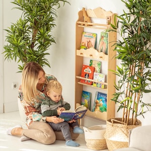 May include: A mother and child sit on the floor reading a book together. A wooden bookshelf with multiple shelves is mounted on the wall behind them. The bookshelf is filled with colorful children's books. The bookshelf has a decorative top with a small wooden giraffe figurine.
