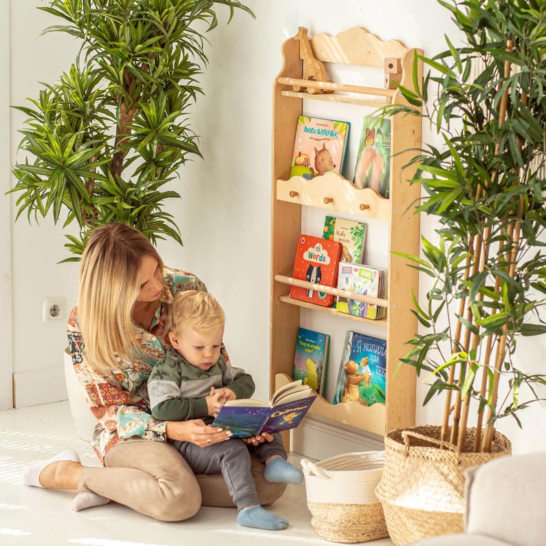 May include: A mother and child sit on the floor reading a book together. A wooden bookshelf with multiple shelves is mounted on the wall behind them. The bookshelf is filled with colorful children's books. The bookshelf has a decorative top with a small wooden giraffe figurine.