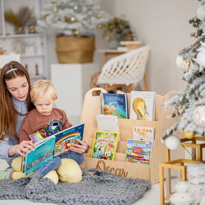 K&ouml;nnte beinhalten: Ein Holz-Buchregal mit einem Griff und dem eingravierten Namen "Oscar" auf der Vorderseite. Das Regal ist mit Kinderb&uuml;chern gef&uuml;llt. Ein junges M&auml;dchen und ein Junge sitzen auf einer grauen Decke und lesen ein Buch zusammen.