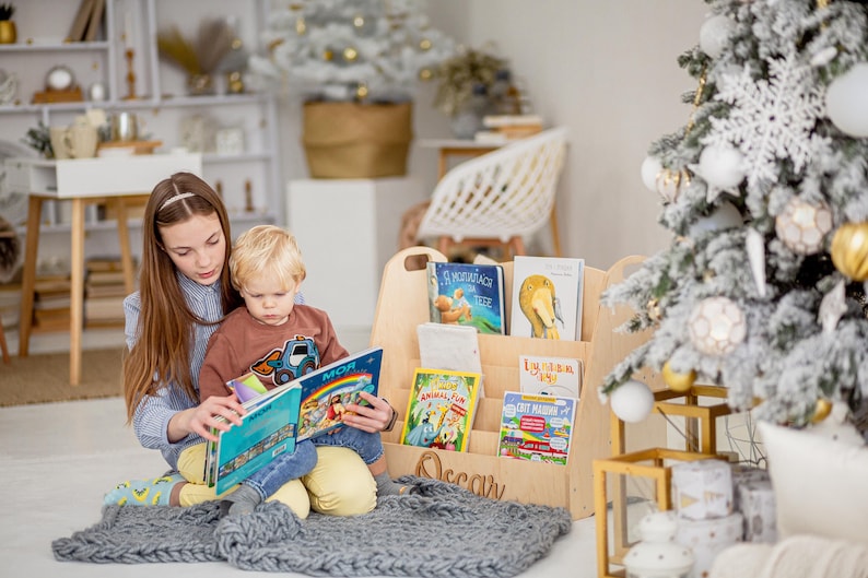 K&ouml;nnte beinhalten: Ein Holz-Buchregal mit einem Griff und dem eingravierten Namen "Oscar" auf der Vorderseite. Das Regal ist mit Kinderb&uuml;chern gef&uuml;llt. Ein junges M&auml;dchen und ein Junge sitzen auf einer grauen Decke und lesen ein Buch zusammen.