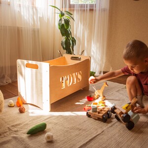 May include: A light-coloured wooden toy box with the word "TOYS" cut out. A child plays with wooden toys, including a giraffe, a car, and blocks, on a neutral rug. The room has a bright, natural aesthetic with a large window.