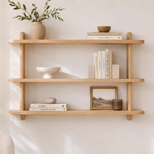 May include: A three-tiered light wood wall shelf featuring rounded edges and vertical supports. The shelves hold a beige vase with greenery, a small wooden bowl, stacked books titled Kinfolk and This Is Home, a stone bowl, and a framed landscape print.