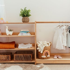 May include: A light wood children's storage unit with shelves and a clothing rack. The shelves hold books, blankets, and woven baskets. The clothing rack displays dresses and shirts. A woven basket with a rainbow decor and a plant in a woven pot are also visible.