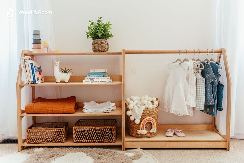May include: A light wood children's storage unit with shelves and a clothing rack. The shelves hold books, blankets, and woven baskets. The clothing rack displays dresses and shirts. A woven basket with a rainbow decor and a plant in a woven pot are also visible.