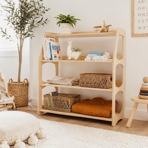 May include: A light-colored wooden bookshelf with four tiers, featuring rounded side supports. The shelves hold books, a white llama-shaped planter, a wooden toy airplane, two woven wicker baskets, a folded beige textile, and a folded orange blanket.