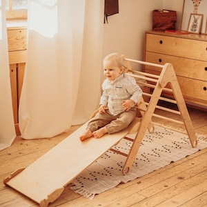 May include: A light-colored wooden climbing triangle with an attached smooth slide ramp. The structure features horizontal rungs for climbing and a flat, angled surface for sliding, constructed from natural wood with a light finish.