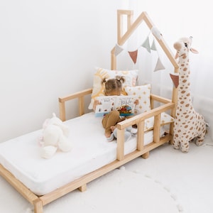 K&ouml;nnte beinhalten: Ein Kinderbett aus Holz mit einem hausf&ouml;rmigen Kopfteil und einer wei&szlig;en Matratze. Ein Kind sitzt auf dem Bett und liest ein Buch mit dem Titel "My Little Bible". Auf dem Bett liegen Stofftiere und Kissen.