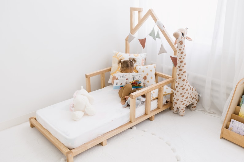 K&ouml;nnte beinhalten: Ein Kinderbett aus Holz mit einem hausf&ouml;rmigen Kopfteil und einer wei&szlig;en Matratze. Ein Kind sitzt auf dem Bett und liest ein Buch mit dem Titel "My Little Bible". Auf dem Bett liegen Stofftiere und Kissen.