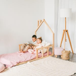 May include: Two children are sitting on a wooden bed frame shaped like a house. The bed has a pink mattress and a white rug underneath. The children are reading a book together.