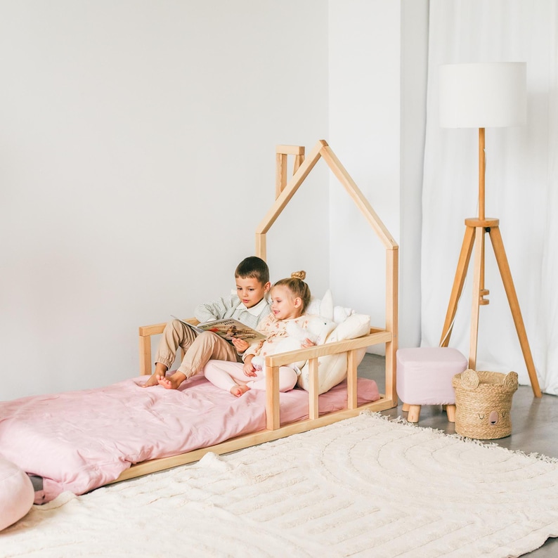 May include: Two children are sitting on a wooden bed frame shaped like a house. The bed has a pink mattress and a white rug underneath. The children are reading a book together.