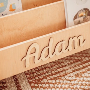 May include: A wooden bookshelf with the name "Adam" written in cursive on the front. The bookshelf has three shelves and is made of light wood.