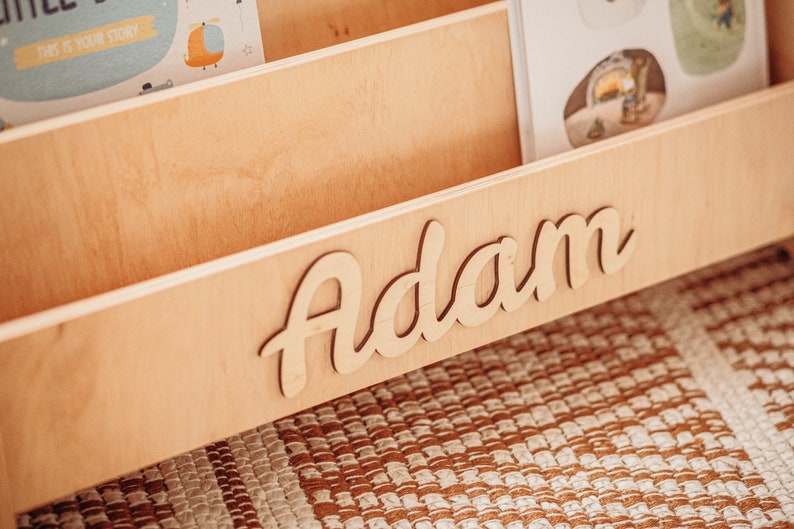 May include: A wooden bookshelf with the name "Adam" written in cursive on the front. The bookshelf has three shelves and is made of light wood.